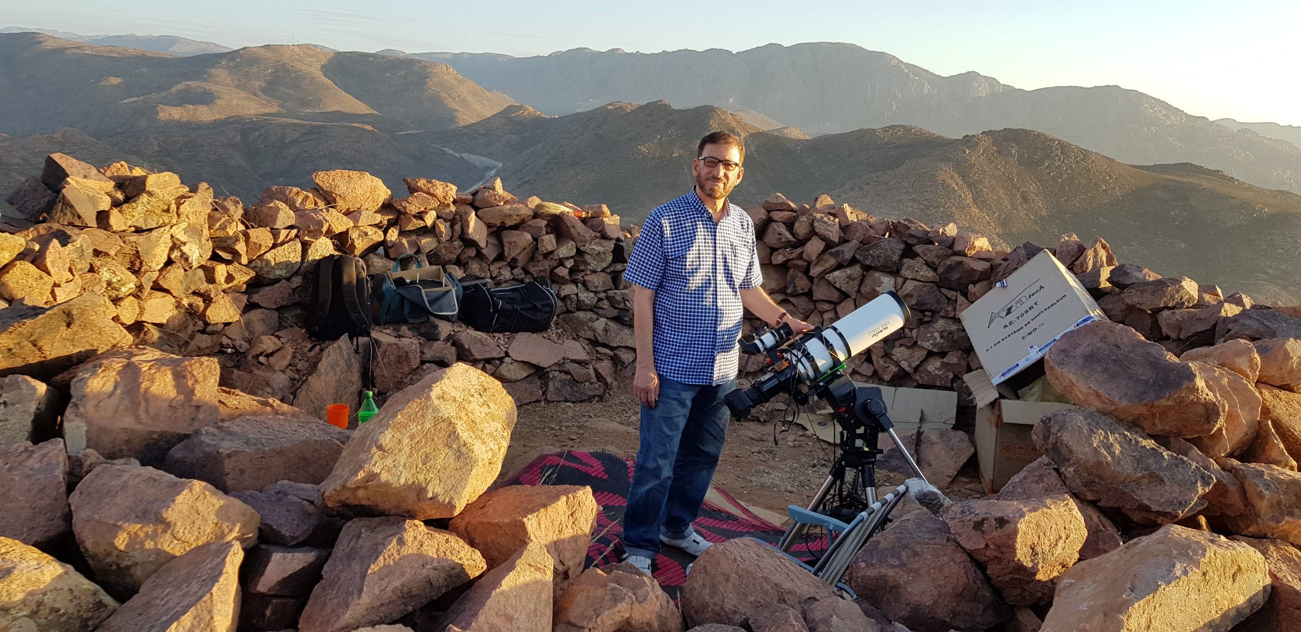 Mazin with his refractor set up amongst the rocks of the summit, illuminated by a low sun, with rugged mountains in the background