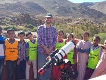 Mazin and ten schoolchildren pose by his telescope in the sun, with rugged mountains in the background