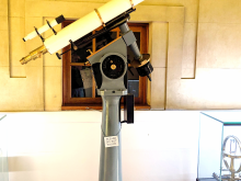 The white telescope tube is mounted on an equatorial mount upon a plinth.
