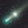The bright nucleus of the greenish comet is at lower left, with the diffuse tail stretching from there to the top-right corner. The dense star cluster appears centrally, behind the tail; a symmetrical sphere of countless stars.