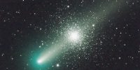 The bright nucleus of the greenish comet is at lower left, with the diffuse tail stretching from there to the top-right corner. The dense star cluster appears centrally, behind the tail; a symmetrical sphere of countless stars.