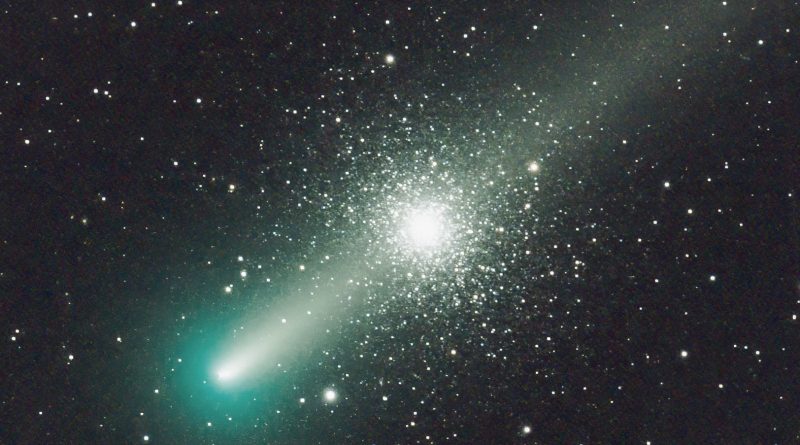 The bright nucleus of the greenish comet is at lower left, with the diffuse tail stretching from there to the top-right corner. The dense star cluster appears centrally, behind the tail; a symmetrical sphere of countless stars.