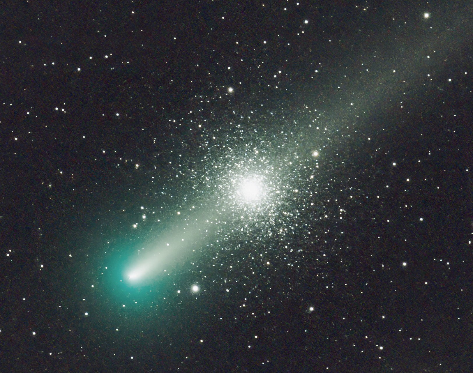 The bright nucleus of the greenish comet is at lower left, with the diffuse tail stretching from there to the top-right corner. The dense star cluster appears centrally, behind the tail; a symmetrical sphere of countless stars.