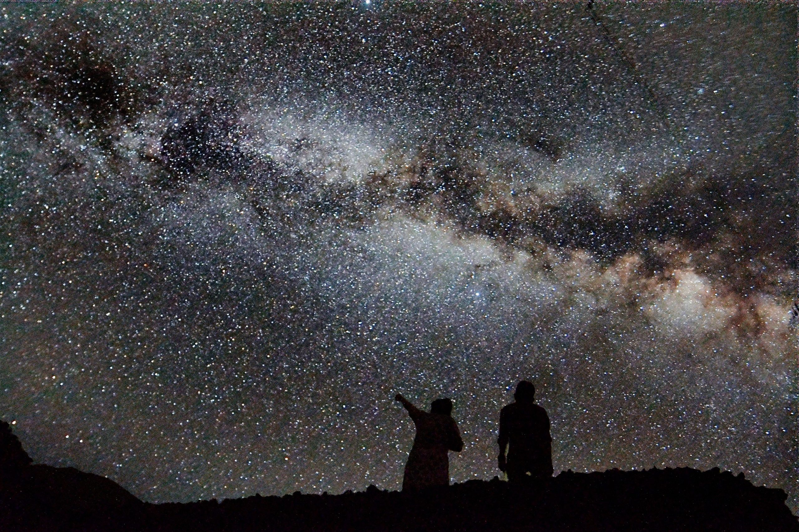 Two figures, one pointing at the sky, appear in silhouette against a densely starry sky with no light pollution, dominated by the Milky Way stretching from left to right with the dark dust lanes prominent