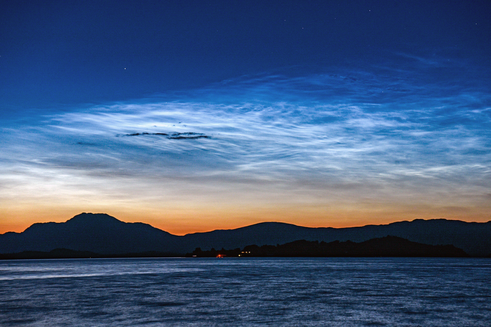 Noctilucent cloud over Loch Lomond. The clouds appear electric blue, bright against a dark sky which graduates to an orange glow above the mountainous horizon. Waves on the Loch's surface are in the foreground.