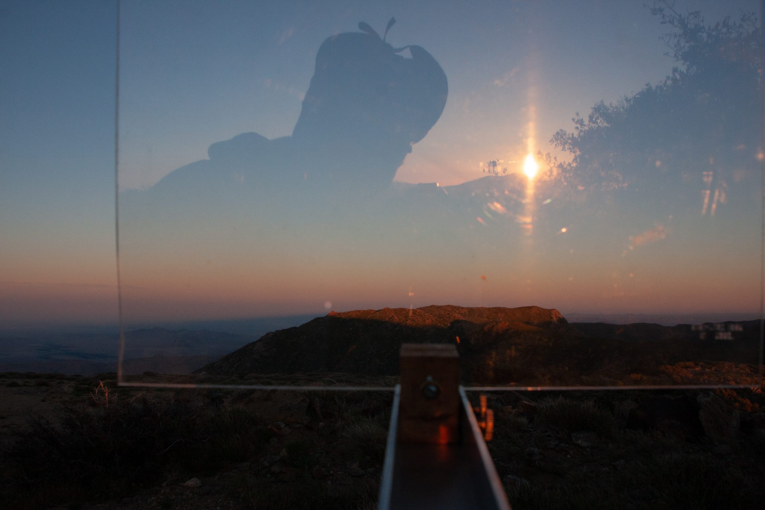 Behind a transparent plastic pane, the Moon can be seen just above a rocky horizon, where a rocky outcrop catches the low sunlight and appears reddish. The Sun is seen reflected in the pane, above and to the right of the Moon. The photographer's silhouette is also seen in the plastic.