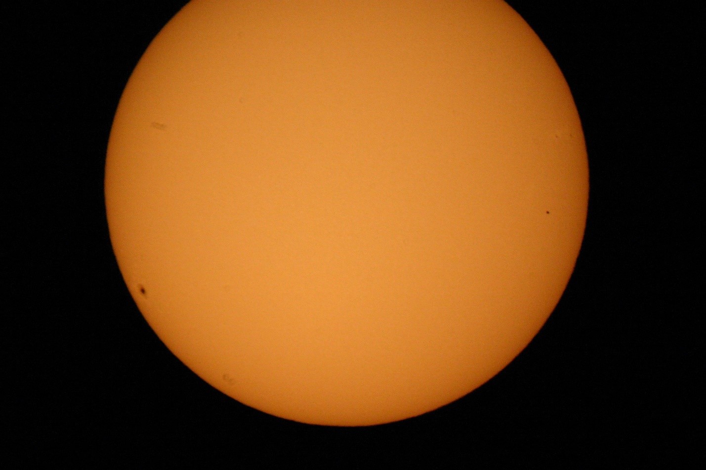 The solar disc: an orange disc against a black background, cut off slightly at the top. Sunspots are visible, especially towards the limb at the left. Mercury is a small black spot, smaller than the sunspot but circular unlike the sunspot's extended (foreshortened) shape