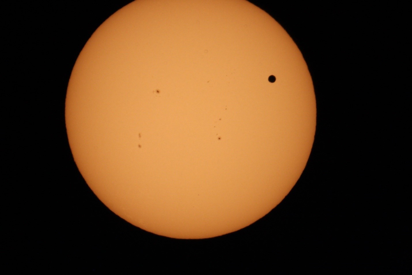 The Sun's photosphere is an orange disc filling most of hte image, against a black background. There are sunspots on the Sun, and a larger circular spot which is Venus.